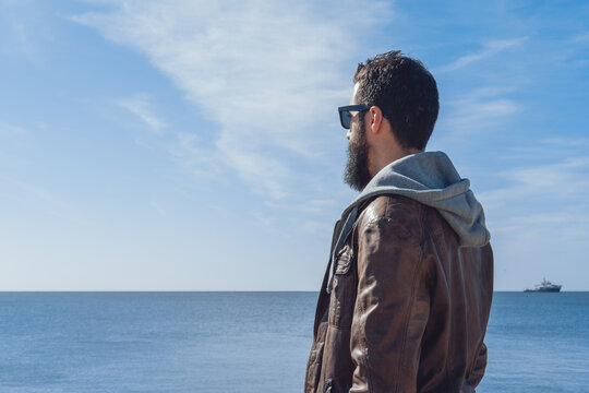 Side view of pensive ethnic bearded male traveler in trendy clothes and sunglasses relaxing on rocky beach of sea in Seven Sisters
