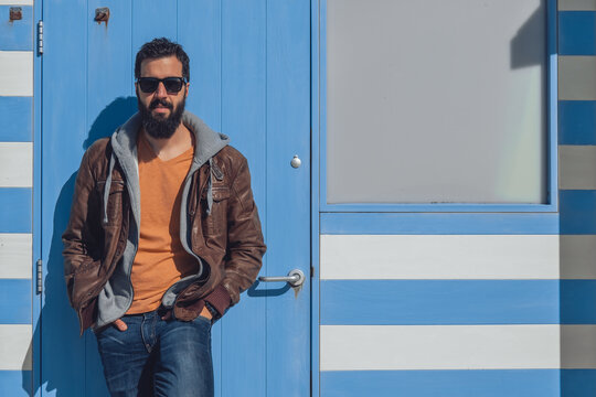 Confident young bearded ethnic male in stylish outfit and sunglasses standing near blue door with hands in pockets while resting in Seven Sister on sunny day