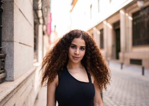 Self assured young ethnic female with long curly hair standing near aged building on street