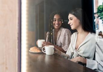 Through window view of cheerful millennial diverse girlfriends chatting happily while sitting at counter in cafe and enjoying hot coffee with pastry
