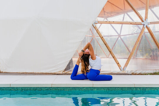 Side view of anonymous barefoot lady sitting in Eka Pada Rajakapotasana pose while reflecting in swimming pool