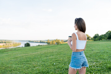 woman in summer park by river, holds cup of coffee and tea in hand, looks into distance, enjoys beautiful view and landscape. Free space for a copy of the text. Background green grass river and lake.