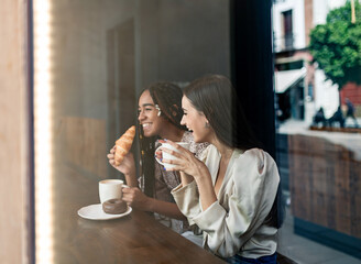 Through window view of cheerful millennial diverse girlfriends chatting happily while sitting at counter in cafe and enjoying hot coffee with pastry