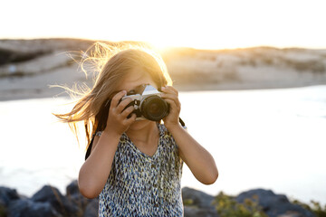 Unrecognizable young female tourist in stylish dress taking photo of sea standing on rocky shore at sunset