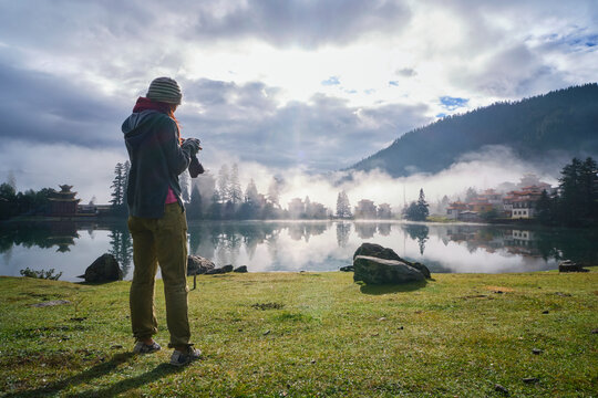 Low angle full body of anonymous tourist with photo camera taking photo of calm lake located in mountainous terrain and oriental shrine