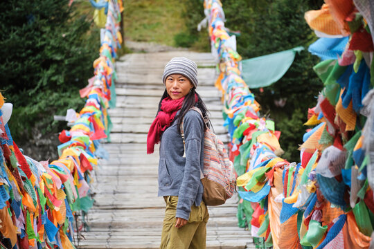 Young ethnic female tourist in warm clothing and hat looking at camera while crossing simple long narrow suspension wooden bridge decorated with bright multi colored pieces of fabric in daylight