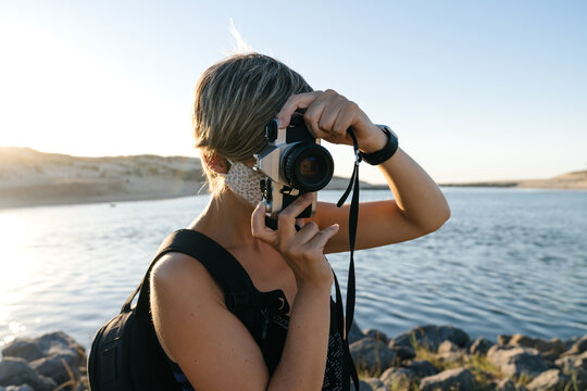 Peaceful adult female tourist with short hair in casual clothes and backpack standing on rocky coast of sea with photo camera wearing medical mask