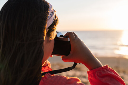 Back view of anonymous stylish teen girl in casual clothes standing on sandy beach and taking photo of sunset over ocean