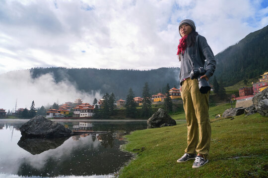 Low angle full body of calm Asian woman with professional photo camera standing on grassy coast of lake against Tibetan Buddhist temple
