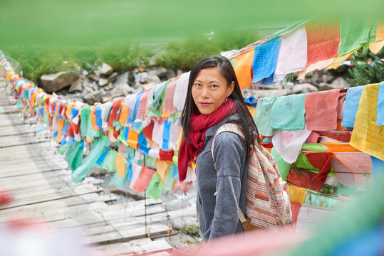 Young ethnic female tourist in warm clothing and hat looking at camera while crossing simple long narrow suspension wooden bridge decorated with bright multi colored pieces of fabric in daylight