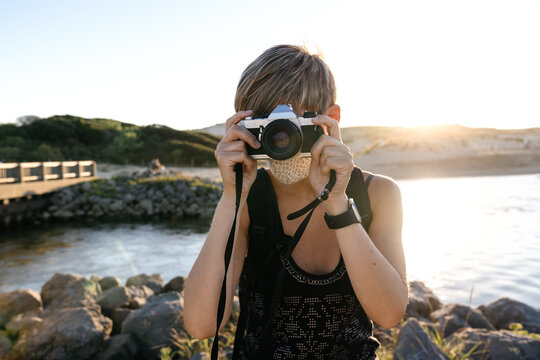 Peaceful adult female tourist with short hair in casual clothes and backpack standing on rocky coast of sea with photo camera wearing medical mask