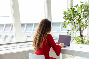 Back view of anonymous female entrepreneur in protective mask sitting at table in workplace and typing on netbook while working on project during coronavirus pandemic