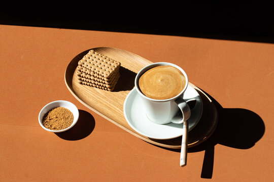 From above of ceramic cup of tasty coffee near crunchy crackers on wooden plate near bowl with cane sugar in cafe in sunlight