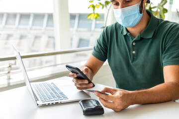 Calm male in protective mask sitting at table with smartphone and plastic card while buying in online app