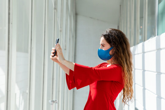 Young woman tourist wearing protective mask standing with suitcase in departure lounge in airport and taking picture with cellphone while waiting for flight during coronavirus
