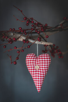 Soft textile checkered heart hanging on tree branch with decorative red berries on gray background in studio