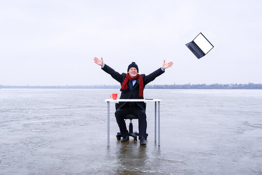 Senior smiling businessman in suit sits with outstretched hands at a table in the middle of a frozen lake in winter and throws away his laptop. Copy space for text on screen of device