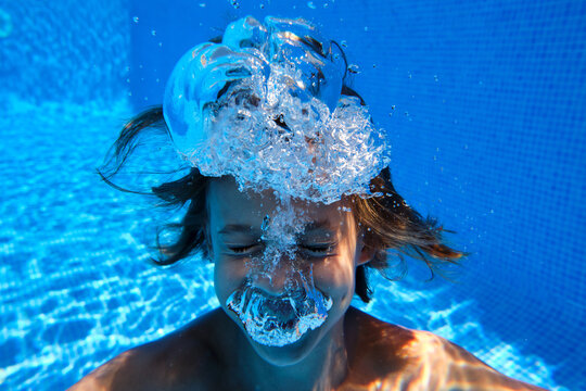Underwater shot of cheerful teenage boy diving into blue swimming pool water with eyes closed and smiling happily