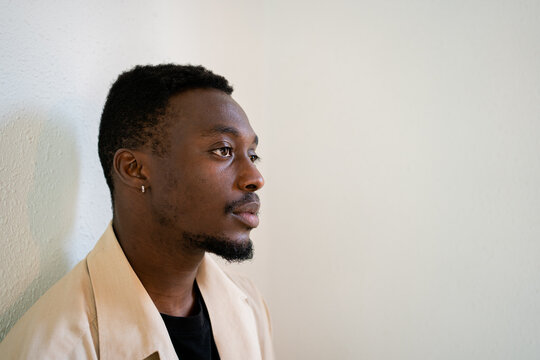 Pensive young African American male in light beige formal shirt thoughtfully looking away while standing near white wall