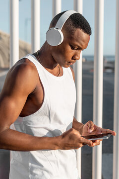 Side view of African American runner browsing mobile phone and listening to music in headphones while training on street