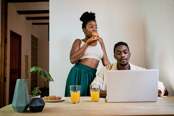 Concentrated black male freelancer working at laptop while thoughtful African American wife in crop top eating bun and looking away