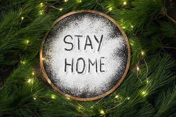 Round dark sign with the words "stay home" in the snow, framed by green pine branches and garland lights