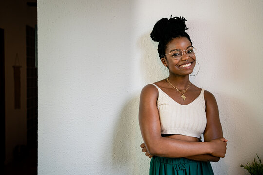 Cheerful African American female in white top and round transparent eyeglasses with dark hair tied up looking at camera while standing in room near grey wall