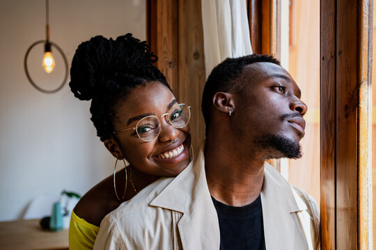 Cheerful young African American female in transparent eyeglasses with bunch looking at thoughtful man standing near window