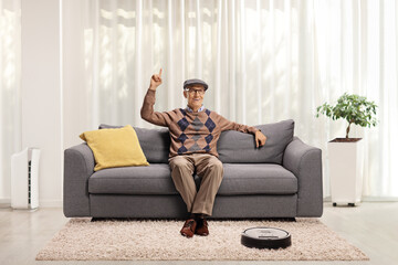 Elderly man sitting on a sofa at home, pointing up and a robotic vacuum cleaner dusting the carpet
