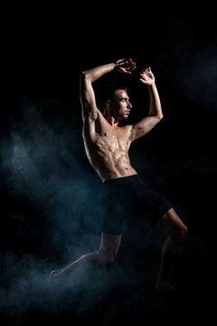 Full body of faceless tanned sporty shirtless dancer in shorts raising arm while doing dance move in dark room in light of lamp looking away