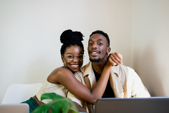 Positive young slim African American wife in casual clothing looking at camera with smile while embracing husband in bright room decorated with green pot plant