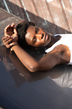 Side view graceful African American female model in white shirt laying back looking at camera on construction on sunny roof and touching head gently