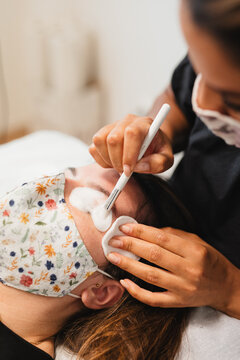 High angle of crop anonymous female master in medical mask applying foaming cleanser on eyelashes of client after extension in modern salon