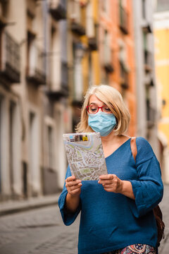 Low angle of female traveler in medical mask for coronavirus prevention holding touristic map and checking route while standing on narrow paved street with old buildings in Cuenca town in Spain