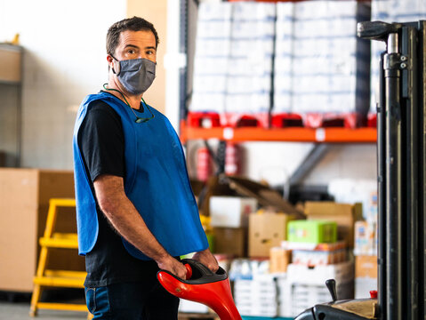Focused adult male worker wearing high visibility vest and face mask holding trolley cart handle and looking at camera while working in contemporary storehouse