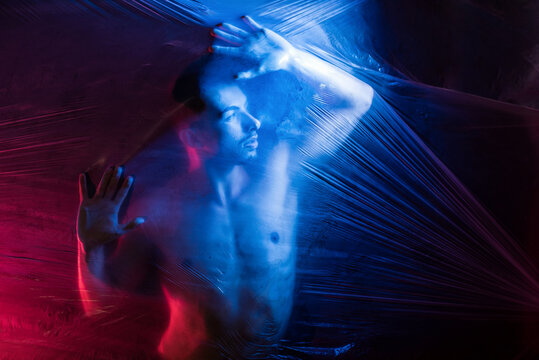 Pensive young handsome shirtless muscular male looking away while putting hands on surface of big transparent tape in light of blue and red lamps