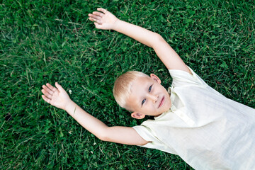 Overhead cheerful blond haired boy in white shirt looking at camera with smile while chilling on verdant grass with arms behind head