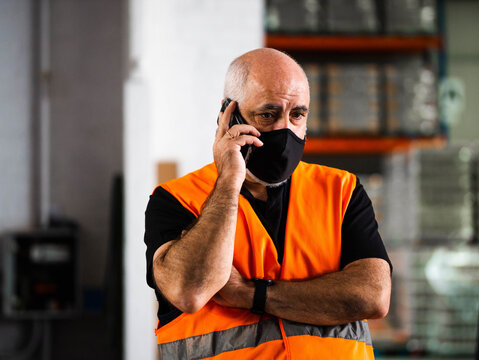 Serious Adult Male Manager In Uniform And Protective Mask Standing With Arms Crossed Near Warehouse Autoloader And Having Conversation On Mobile Phone