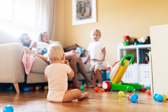 Back view of baby sitting on couch with toys spending time with brother and parents in living room