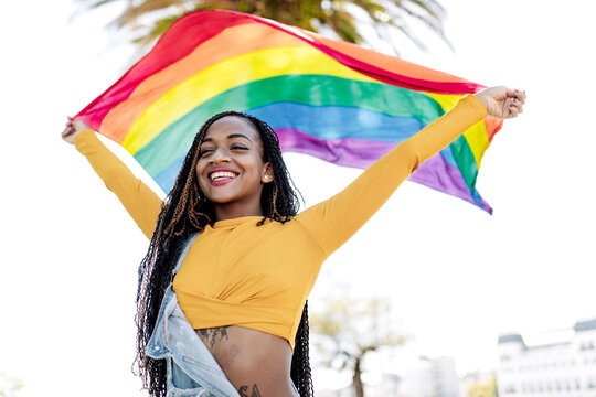 Low angle of proud African American female standing with fluttering LGBT rainbow flag on street and cheerfully looking away