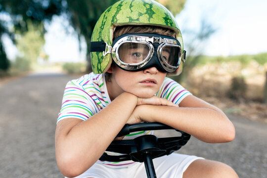 Crop dreamy boy in safety glasses and decorative helmet sitting leaned with hands on steering wheel on road while looking away