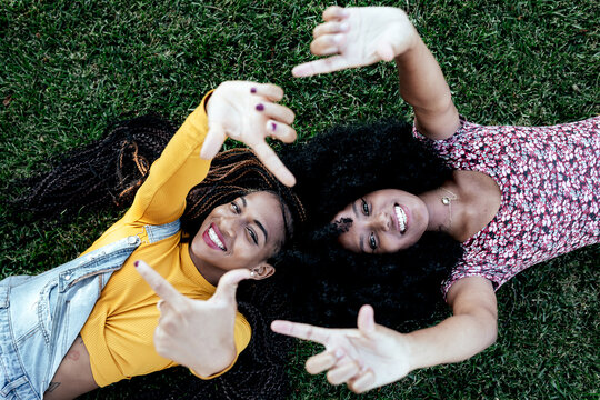 Top view of cheerful African American female friends lying on green lawn in park and looking at camera while spending weekend in summer together
