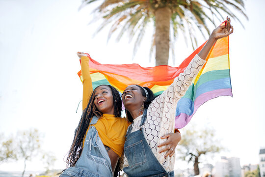 Low angle of delighted African American females in love hugging on street while standing with LGBT rainbow flag and looking away