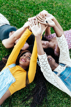 High angle of happy multiracial girlfriends lying together on grass and stacking hands while resting in park in summer