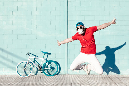 Expressive young bearded male hipster in trendy outfit and medical mask jumping with outstretched arms and screaming near bicycle leaning on blue wall on street
