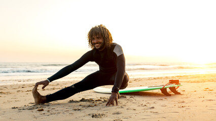 Full body side view of African American surfer with curly hair in wet suit stretching leg on sandy beach of sea at sunset