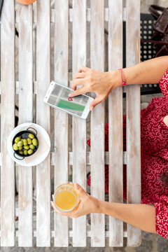 Top view of crop unrecognizable female with glass of refreshing drink touching screen on cellphone with map application while sitting at table with bowl of olives