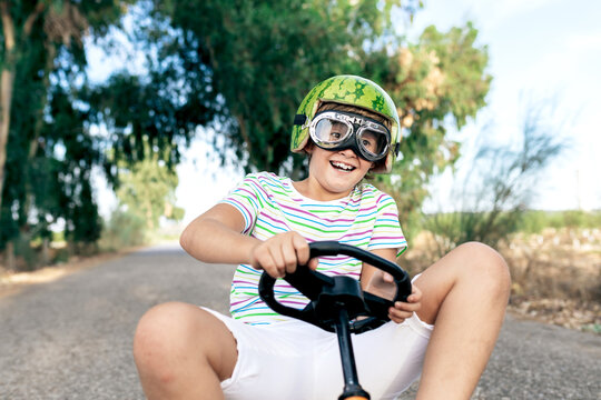 Low angle of amazed kid in trendy wear and goggles riding go kart with pedals on roadway while looking away