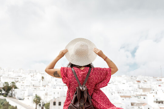 Back view of anonymous stylish tourist with straw hat and raised arms contemplating old houses in Rhodes under cloudy sky