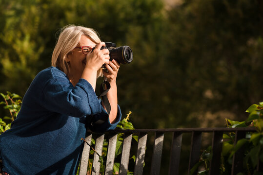 Side view of serious adult blond female photographer with professional photo camera leaning on fence and looking away while standing against green bushes and taking pictures of nature